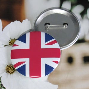 Puede incluir: Un botón redondo con un diseño de bandera Union Jack rojo, blanco y azul. El botón tiene un diámetro de aproximadamente 5 cm y una parte posterior de metal plateado.
