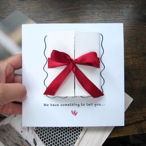 May include: A square white greeting card featuring a central gatefold design secured by a deep red satin ribbon tied in a bow. The card includes a black wavy border outline, the text "We have something to tell you..." and two small pink hearts.