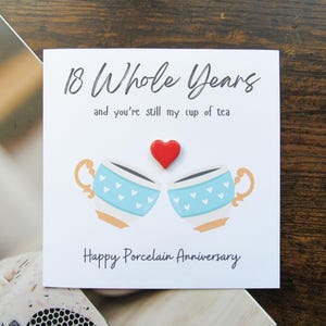 以下が含まれることがあります： 「18 Whole Years and you're still my cup of tea」と書かれた白いグリーティングカード。赤いハートの下に、ハートのディテールが施された2つの青いティーカップが描かれています。カードには「Happy Porcelain Anniversary」と書かれています。