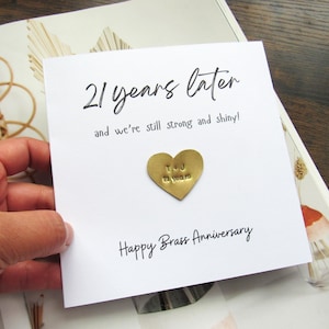 May include: A white card with the text "21 years later" and "and we're still strong and shiny!" in black script. A gold heart-shaped charm is affixed to the card, with the initials "T & J" and "21 years" stamped on it. The card reads "Happy Brass Anniversary".