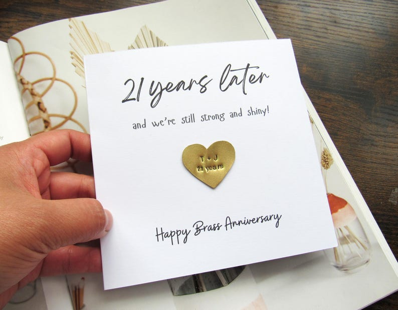 May include: A white card with the text "21 years later" and "and we're still strong and shiny!" in black script. A gold heart-shaped charm is affixed to the card, with the initials "T & J" and "21 years" stamped on it. The card reads "Happy Brass Anniversary".