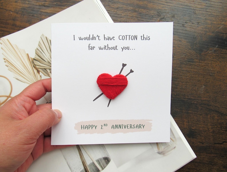 May include: A white anniversary card with the text "I wouldn't have COTTON this far without you..." and "HAPPY 2nd ANNIVERSARY". The card features a red yarn heart pierced by two black needles.