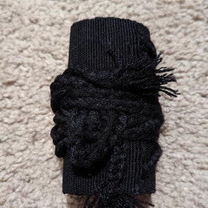 May include: A black fabric cuff with a textured, woven design. The cuff is wrapped with a decorative, knotted element and finished with fringed edges. The item is likely a decorative accessory.