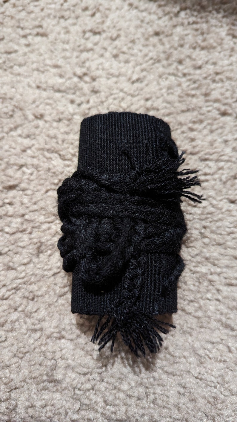 May include: A black fabric cuff with a textured, woven design. The cuff is wrapped with a decorative, knotted element and finished with fringed edges. The item is likely a decorative accessory.