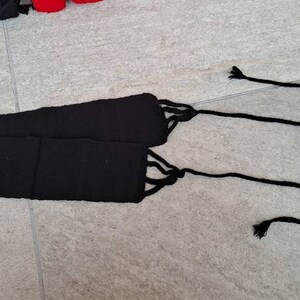 May include: A black woven belt with long, knotted ties at each end. The belt is made of a thick, textured material and is lying on a gray tiled surface. The ties are thin and hang down.