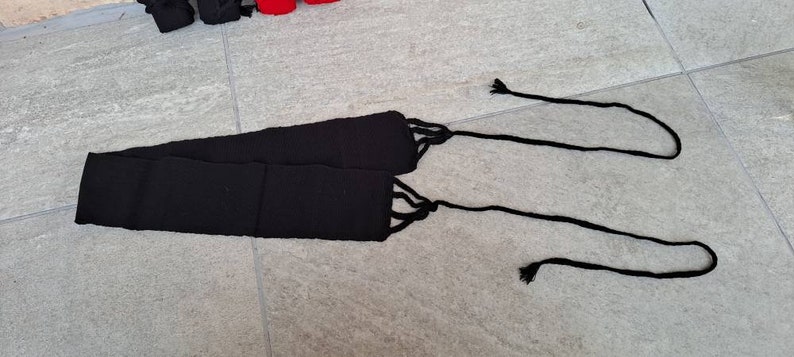 May include: A black woven belt with long, knotted ties at each end. The belt is made of a thick, textured material and is lying on a gray tiled surface. The ties are thin and hang down.