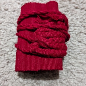 May include: A rectangular, deep red fabric piece wrapped with thick, matching red yarn. The yarn is looped and knotted around the fabric, creating a textured, decorative effect. The item is likely a craft supply or embellishment.
