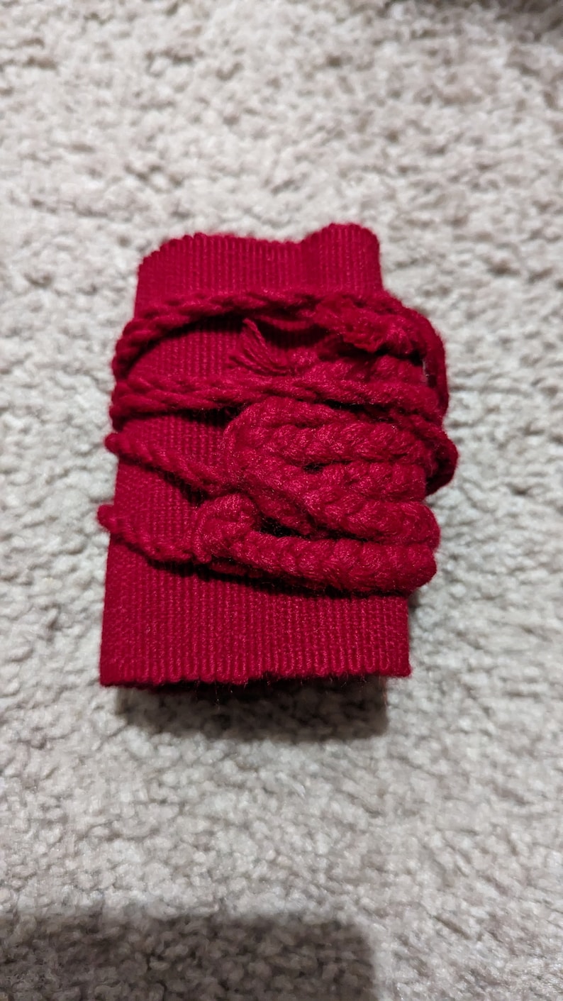 May include: A rectangular, deep red fabric piece wrapped with thick, matching red yarn. The yarn is looped and knotted around the fabric, creating a textured, decorative effect. The item is likely a craft supply or embellishment.