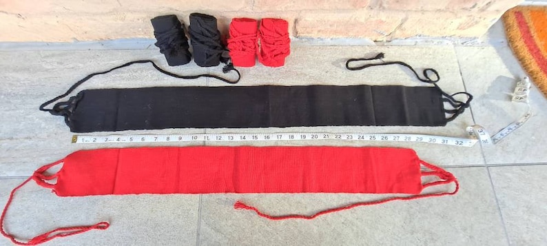 May include: A flat lay of black and red martial arts equipment. Two black and two red hand wraps are at the top. Below are two belts, one black and one red, with a measuring tape alongside.