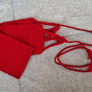 May include: A vibrant red sash with a rectangular body and braided cord ties. The sash is made of a textured fabric and is displayed on a light gray tiled surface. The cord ends in a small tassel.