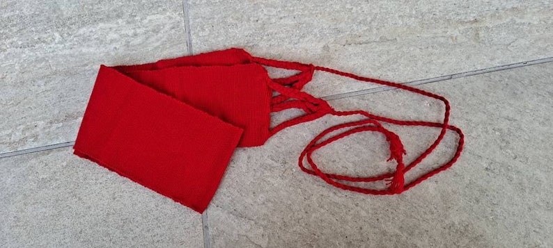 May include: A vibrant red sash with a rectangular body and braided cord ties. The sash is made of a textured fabric and is displayed on a light gray tiled surface. The cord ends in a small tassel.