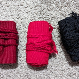 May include: Three fabric wrist wraps in red and black. Two red wraps are wrapped with red rope, and one black wrap is wrapped with black rope. The wraps are on a light gray, textured surface.
