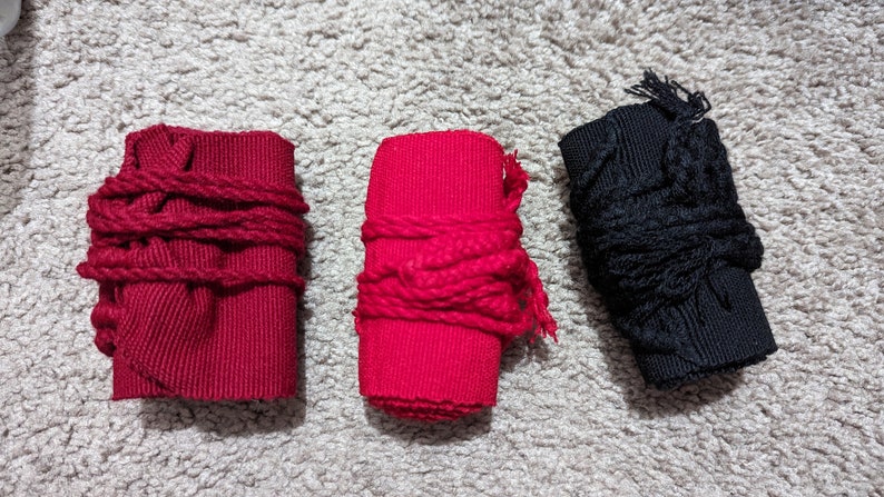 May include: Three fabric wrist wraps in red and black. Two red wraps are wrapped with red rope, and one black wrap is wrapped with black rope. The wraps are on a light gray, textured surface.