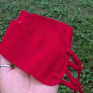 May include: Bright red fabric wristbands with braided ties. The wristbands are made of a ribbed material and are folded over. The background is green grass.