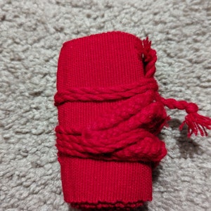 May include: A cylindrical, bright red fabric object wrapped with a thicker, twisted red cord. The cord is tied around the cylinder and has a tassel at the end. The object is on a gray, textured surface.