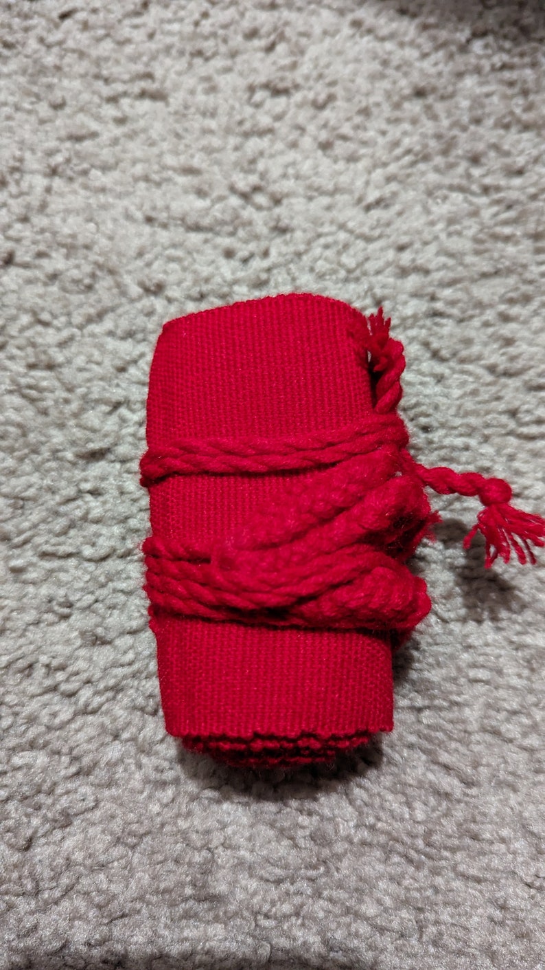 May include: A cylindrical, bright red fabric object wrapped with a thicker, twisted red cord. The cord is tied around the cylinder and has a tassel at the end. The object is on a gray, textured surface.
