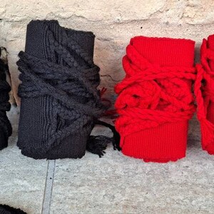 May include: Four wrist wraps, two black and two red, are displayed against a textured, light-colored wall. The black wraps are wrapped with black rope, while the red wraps are wrapped with red rope. The wraps are made of a ribbed fabric.