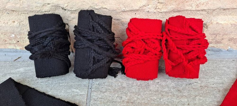 May include: Four wrist wraps, two black and two red, are displayed against a textured, light-colored wall. The black wraps are wrapped with black rope, while the red wraps are wrapped with red rope. The wraps are made of a ribbed fabric.