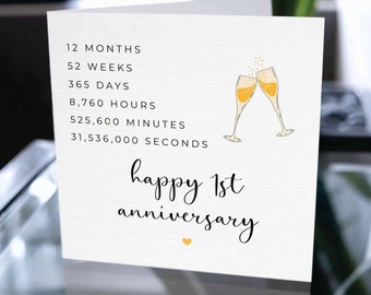 May include: A white anniversary card with the text "happy 1st anniversary" in cursive script. The card includes a list of time increments and a graphic of two champagne glasses clinking together. A small yellow heart is at the bottom.