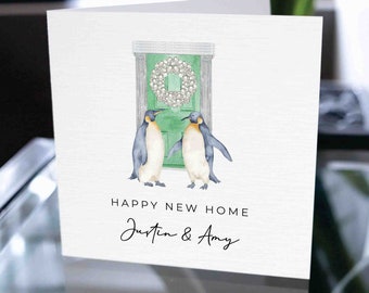 Puede incluir: Una tarjeta blanca con una puerta verde adornada con una corona de perlas blancas. Dos pingüinos se paran frente a la puerta. El texto "Happy New Home Justin & Amy" está escrito debajo de los pingüinos.