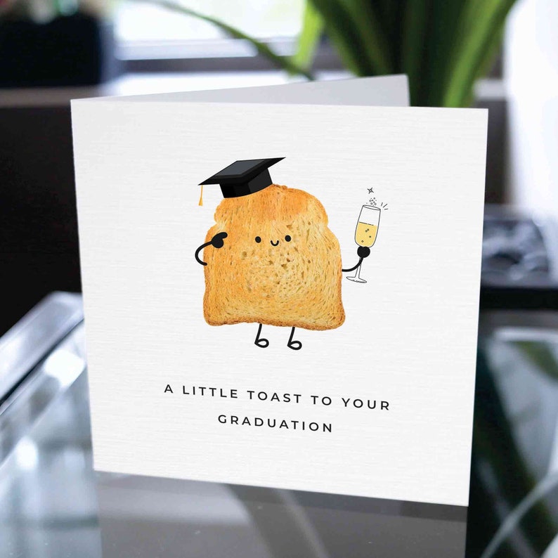 May include: A white card with a cartoon illustration of a slice of toast wearing a graduation cap and holding a glass of champagne. The text "A little toast to your graduation" is printed below the illustration.