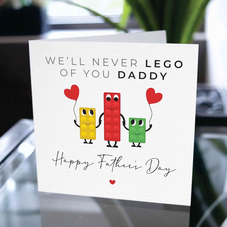 K&ouml;nnte beinhalten: Eine Vatertagskarte mit Lego-Motiv. Sie zeigt drei Lego-Bl&ouml;cke, die sich an den H&auml;nden halten und rote Herzen an Ballons haben. Die Karte lautet "We'll never Lego of you Daddy" und "Happy Father's Day".