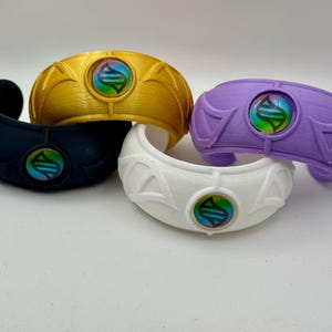 May include: Four cuff bracelets in black, gold, white, and purple. Each bracelet features a central circular emblem with a holographic, iridescent finish displaying a stylized DNA-like symbol. The bands have a raised, geometric wing-like design.