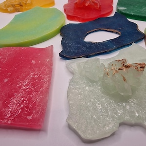 Puede incluir: Una colección de piezas de resina coloridas de forma irregular. Las piezas varían en color, incluyendo rojo, azul, verde, amarillo y un verde claro con degradado amarillo. Algunas piezas tienen elementos decorativos incrustados y todas tienen un acabado brillante.