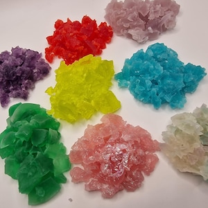 Könnte beinhalten: Eine Auswahl an farbenfrohen, kristallinen Formationen in Rot, Lila, Gelb, Blau, Grün, Rosa und einer Mischung aus Weiß und Grün. Die Kristalle sind zusammengefasst und erzeugen einen strukturierten Eindruck vor weißem Hintergrund.