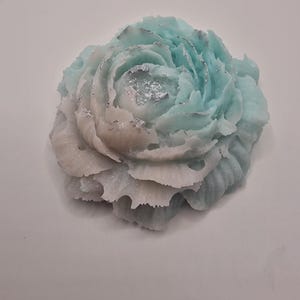 May include: A flower-shaped bar of soap featuring a gradient of colours transitioning from light blue at the top to white at the base. The petals are textured with ruffled edges and accented with small, shimmering silver flecks throughout the surface.