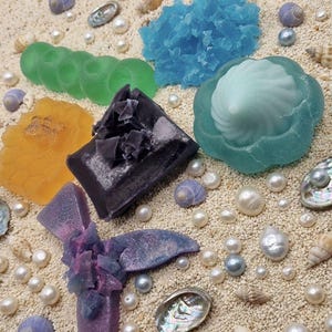 Puede incluir: Una variedad de jabones decorativos coloridos en varias formas, incluyendo una flor y una barra, dispuestos sobre una superficie arenosa con conchas marinas y perlas. Los jabones son en tonos de azul, verde, naranja y morado.