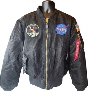 May include: Black bomber jacket with gold zipper and multiple patches, including the NASA logo, an American flag, and an Apollo 11 patch. The jacket has a red "REMOVE BEFORE FLIGHT" tag attached to the zipper.