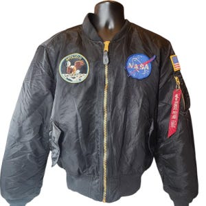 May include: A black bomber jacket featuring a gold-tone front zipper, a ribbed collar, and ribbed cuffs. The chest displays an Apollo 11 mission patch and a blue NASA logo patch. A small American flag patch and a red tag reading Remove Before Flight.