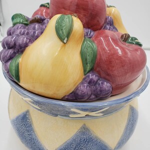 May include: Ceramic cookie jar with a blue and yellow geometric pattern. The lid is shaped like a fruit basket with a pear, apples, and grapes.