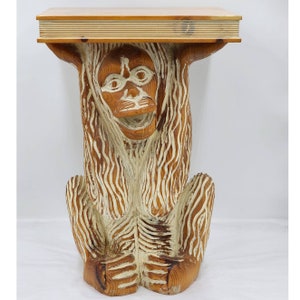 May include: A wooden table with a carved monkey head and body. The table is made of light brown wood with white painted details. The monkey's face is painted with white eyes and a white mouth.