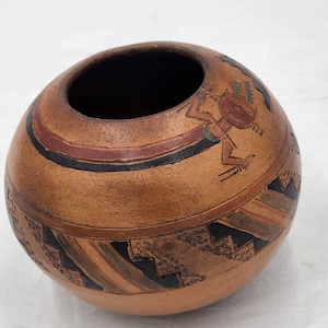 May include: A small, round, terracotta pot with a geometric design painted in black, red, and white. The pot features a stylized figure of a person with a headdress and a staff.