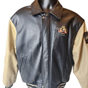 May include: Black and tan leather jacket with a black collar and tan sleeves. The jacket features a Disneyland patch on the chest and a striped hem. The jacket is displayed on a black mannequin.