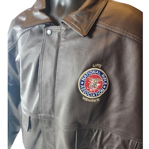 May include: A brown leather jacket with a collar and button closure. The jacket features a patch with the text "National Rifle Association" and an eagle emblem. The jacket has two front pockets.