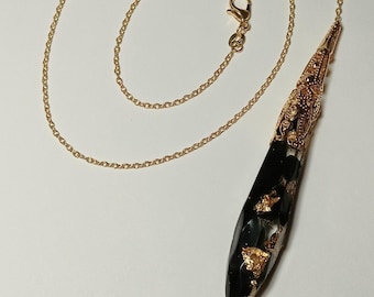May include: A gold-colored chain necklace with a pendant. The pendant is black with gold flakes and a gold-colored filigree cap. The necklace is on a white background.