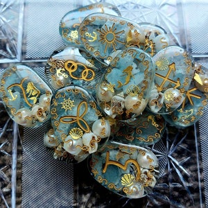 May include: A collection of blue and gold resin charms with nautical themes. Each charm features a different design, including anchors, mermaids, seashells, and compasses. The charms are decorated with gold glitter and white pearl-like accents.