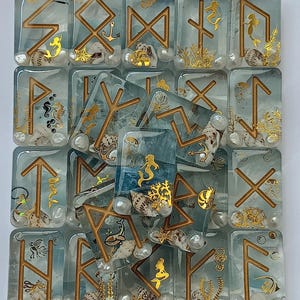 May include: A set of 16 resin tiles featuring Norse runes and ocean-themed imagery. Each tile has a different rune and a variety of sea-life designs, including mermaids, anchors, and seashells. The tiles are a light blue color with gold accents.