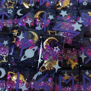 May include: A collection of small, square resin dice with purple and gold glitter embedded in the resin. The dice feature a variety of symbols, including stars, moons, and runes. The dice are perfect for tabletop gaming or as decorative accents.