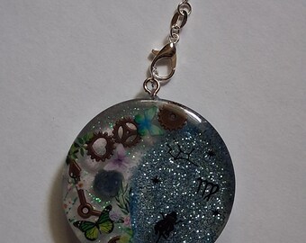 May include: A round pendant with a silver chain and clasp. The pendant is split into two sections. One side features gears, keys, butterflies, and flowers. The other side has a glittery blue background with a silhouette and text.