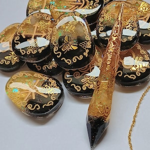 May include: A collection of black and gold resin cabochons with bee and geometric designs, and a black and gold crystal pendulum on a gold chain.