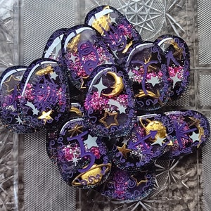May include: A set of 10 purple and gold resin heart-shaped charms with glitter and star designs. Each charm features a different symbol, including a crescent moon, a star, and a wand.