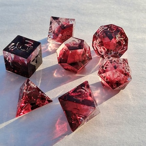 Può includere: Set di sette dadi poliedrici con base in resina trasparente e interno rosso scuro a vortice. I dadi hanno varie forme, tra cui un cubo, un d20 e un d4, con numerazione bianca. Ideali per giochi da tavolo.