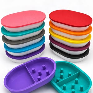 May include: A collection of oval-shaped soap dishes in various colors including pink, teal, gray, blue, purple, red, orange, yellow, and black. Each dish features a white base and an interior divider with small vertical pegs for drainage.