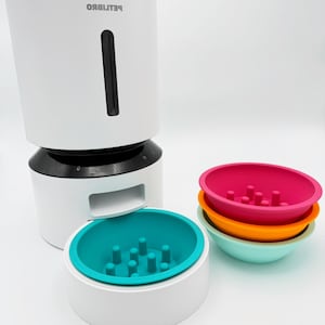 Peut inclure: Un distributeur automatique de nourriture pour animaux de compagnie blanc et noir avec une fenêtre transparente et le mot "PETLIBRO" imprimé sur le devant. Comprend un bol d'alimentation lente turquoise et une pile de trois bols colorés : rose, orange et turquoise.