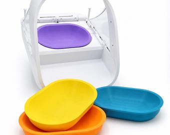 May include: A white automated pet feeder with a purple bowl insert, accompanied by three additional removable oval bowls in bright yellow, orange, and blue. The feeder features a curved white frame and a clear plastic cover over the feeding area.