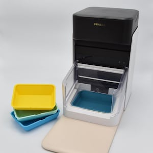 Puede incluir: Un comedero automático para mascotas con tapa negra y cuerpo blanco. El comedero tiene una bandeja transparente y un cuenco azul. Tres bandejas de comida coloridas en amarillo, verde y azul están apiladas a la izquierda. La marca "PETLIBRO" es visible.